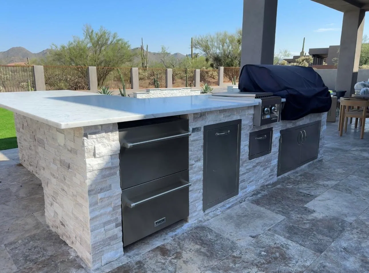 Outdoor kitchen island with stacked stone base and stainless grill on a travertine patio
